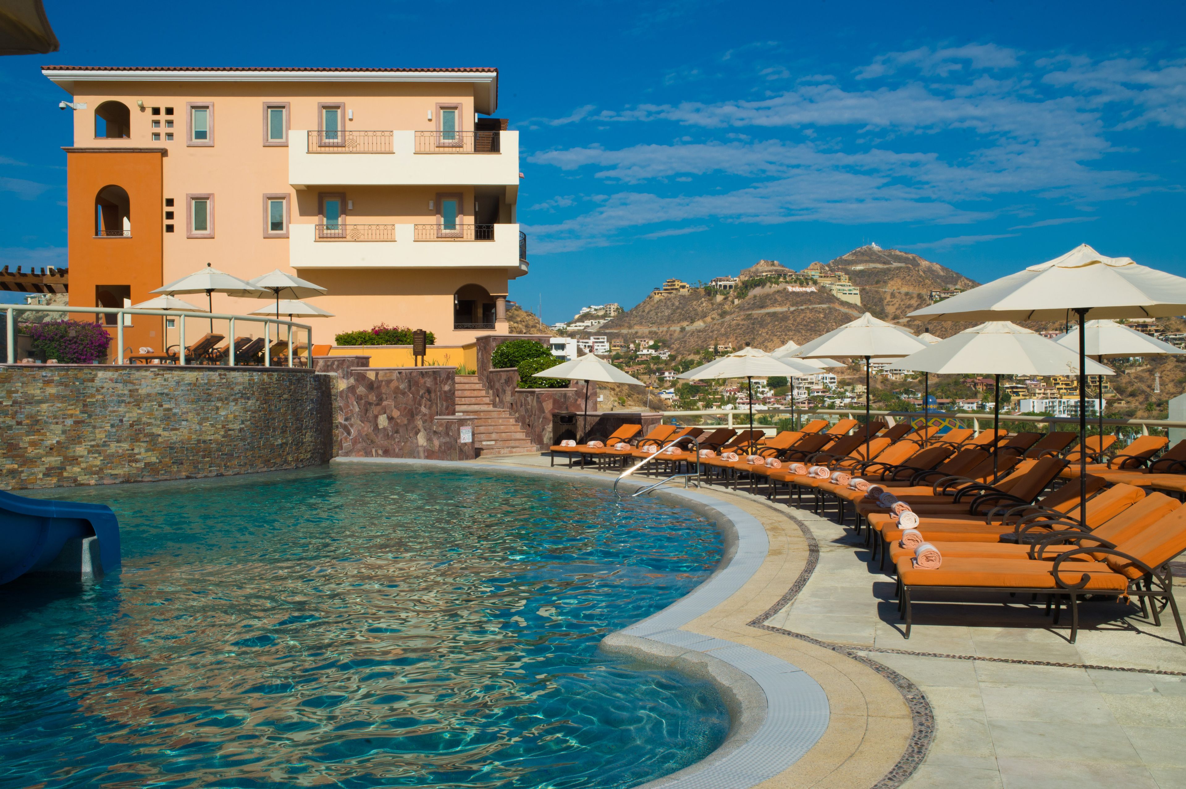A pool area with lounge chairs and umbrellas, a building with balconies, and a mountainous backdrop.