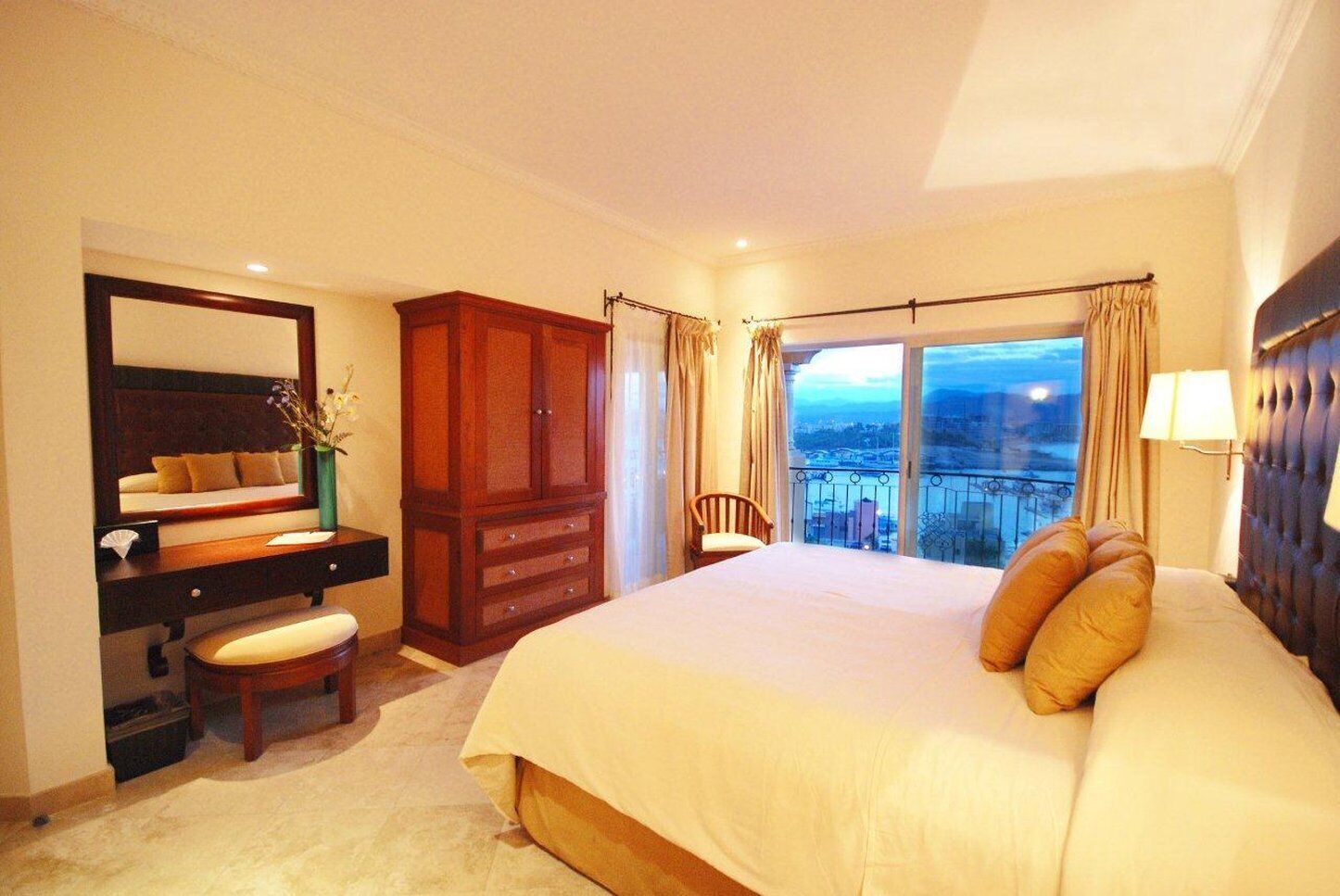 Une chambre d’hôtel avec un grand lit, une coiffeuse avec un miroir, une armoire en bois, une chaise et un balcon avec vue.