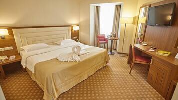 A hotel room with a large bed, a desk with a TV, a chair, and a small table with a lamp.