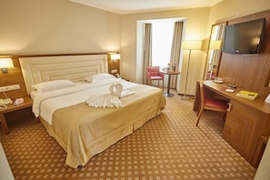 Habitación de hotel con una cama grande, un escritorio con televisor, una silla y una mesita de noche con lámpara.