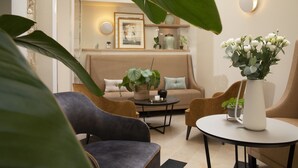A modern living room with a sofa, two armchairs, a coffee table, and a side table. There is a large plant in the foreground, a framed picture on the wall, and a vase with flowers on the side table. - Hotel Des Champs Elysees (Paris)