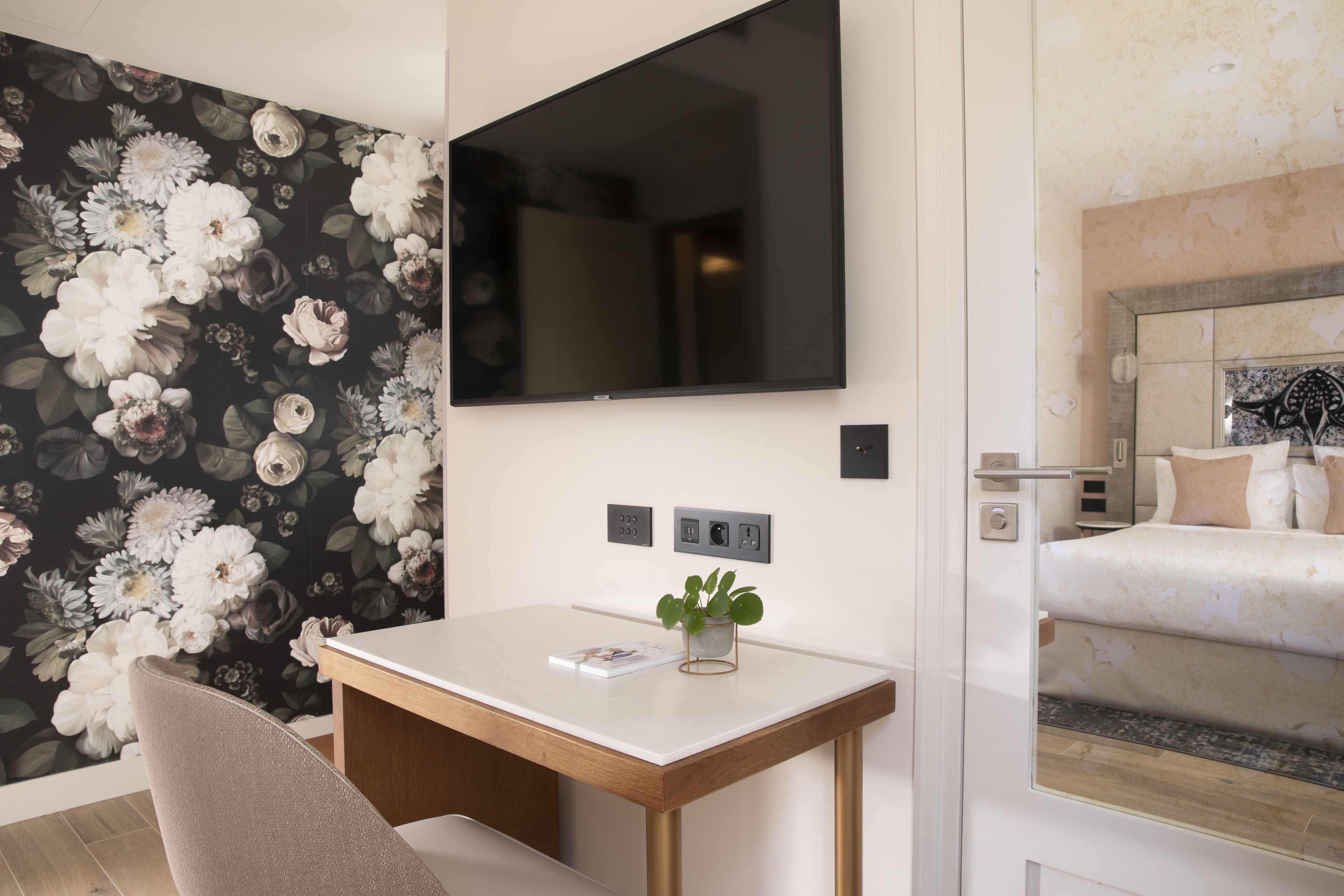 A hotel room with a bed, a TV mounted on the wall, a desk with a plant, and a floral wallpaper.