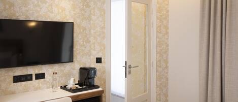 A hotel room with a flat-screen TV mounted on the wall, a desk with a coffee maker, a glass of water, and a tray with a cup and a book.