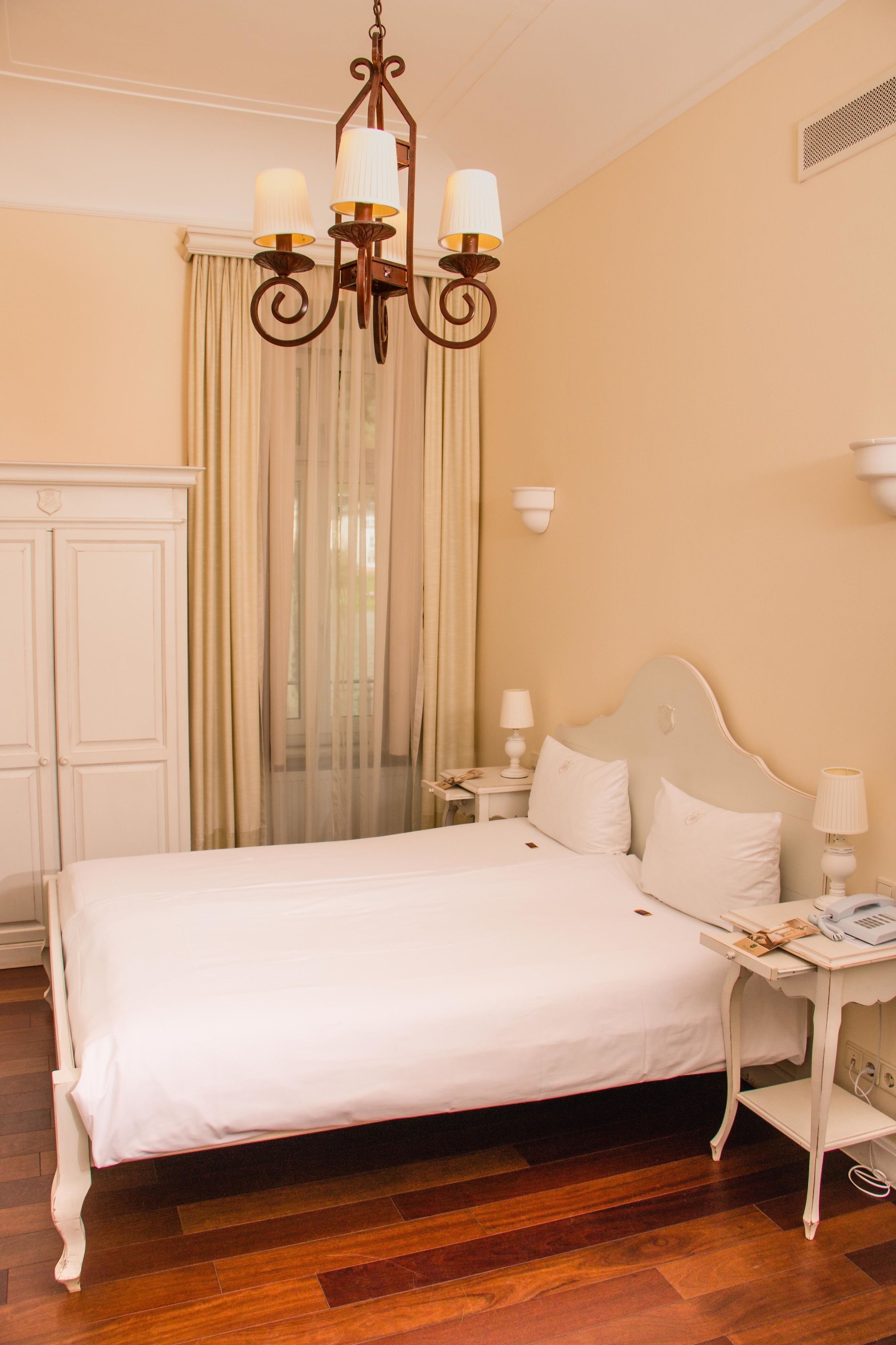 Una habitación de hotel con una cama grande, una araña de cristal, una mesita de noche con lámpara, un armario y una ventana con cortinas.
