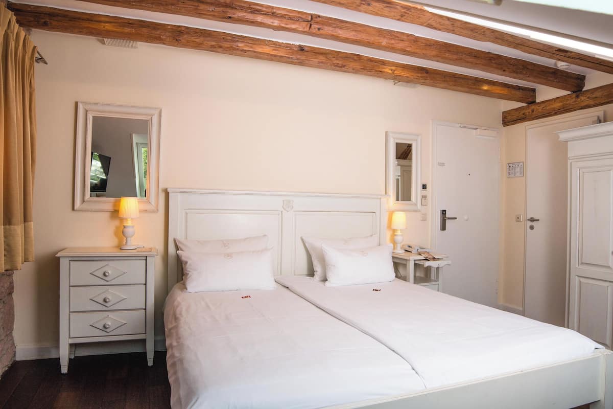 A bedroom with two beds, wooden beams on the ceiling, a nightstand with a lamp, and a mirror on the wall.