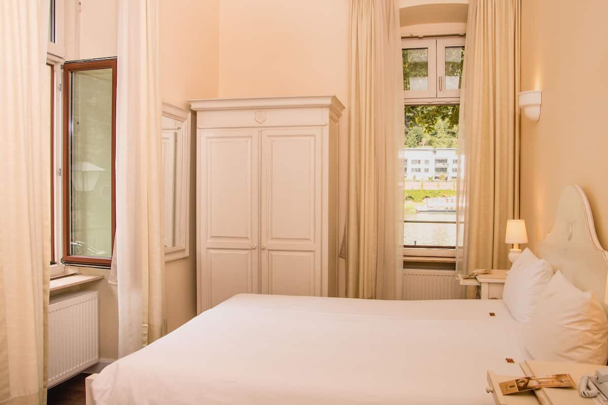 A hotel room with a large bed, a wardrobe, a window with curtains, and a bedside table with a lamp.