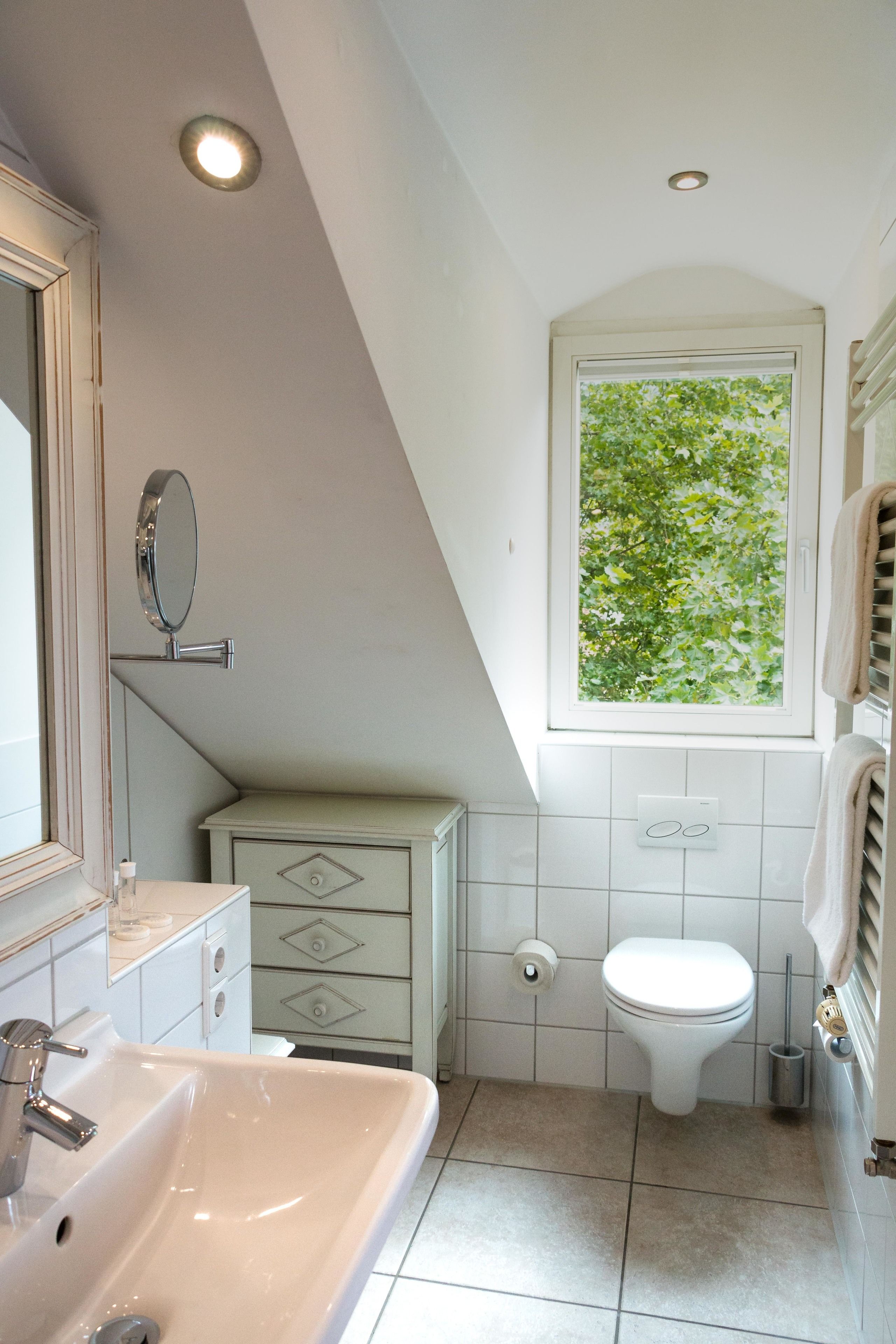 Une salle de bain avec un lavabo blanc, des toilettes, une fenêtre donnant sur des arbres et un miroir.