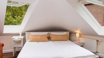 Quarto com teto inclinado, duas janelas, cama com roupa de cama branca e mesa de cabeceira com abajur.