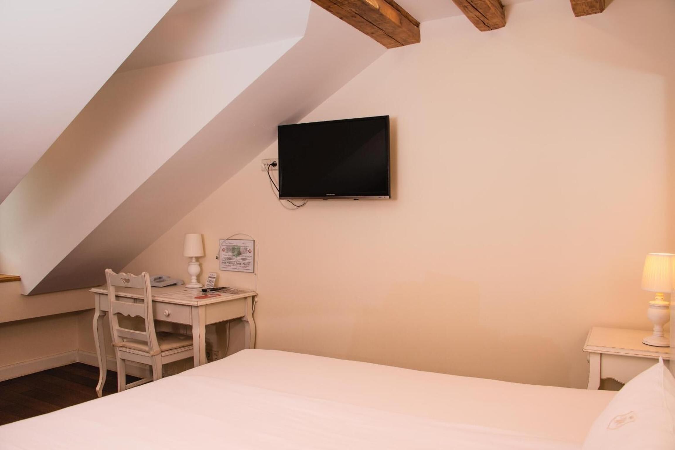 A bedroom with a bed, a desk, a chair, a TV mounted on the wall, and a nightstand with a lamp.