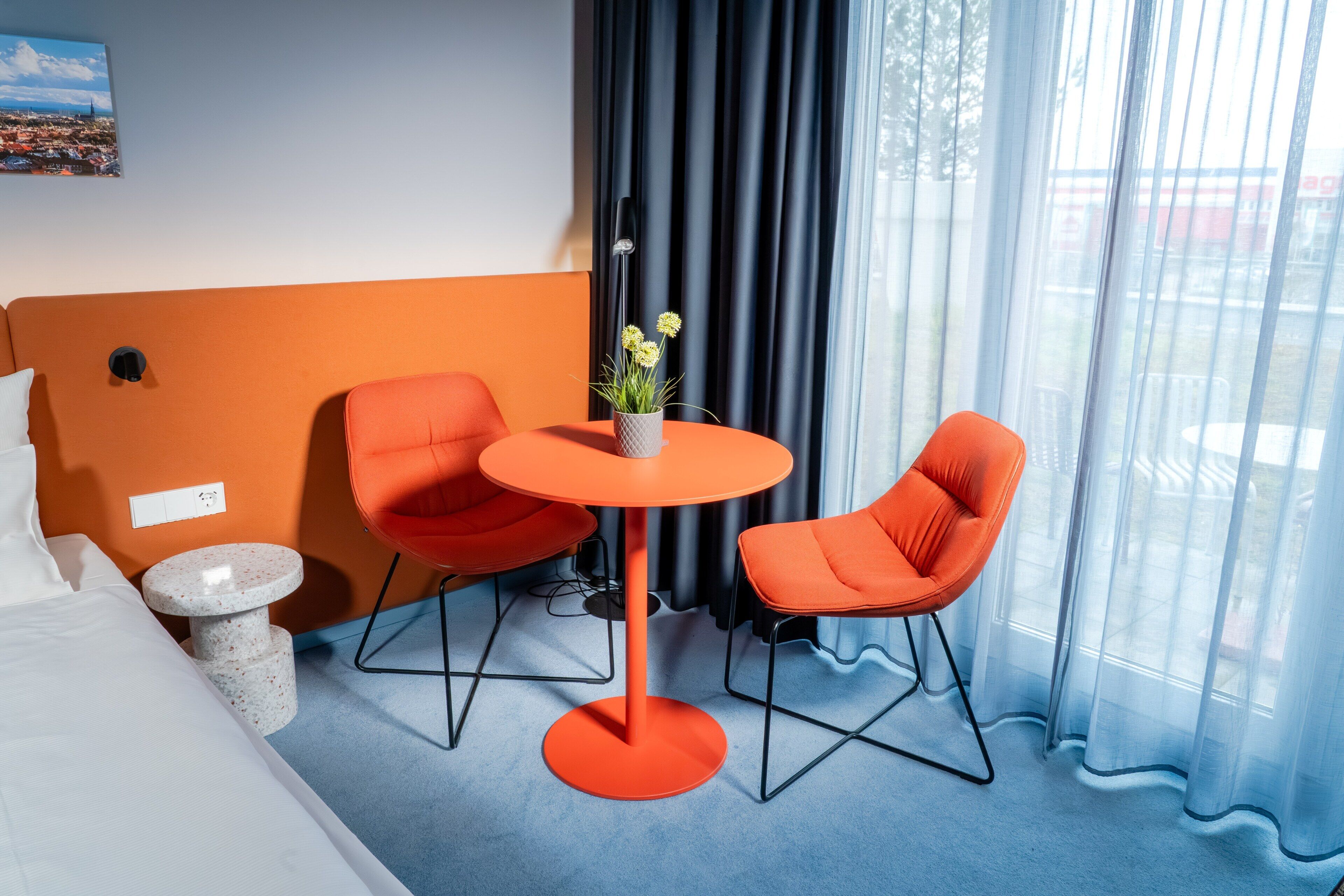 A hotel room with a bed, two orange chairs, a small table with a plant, a painting on the wall, and a window with curtains.