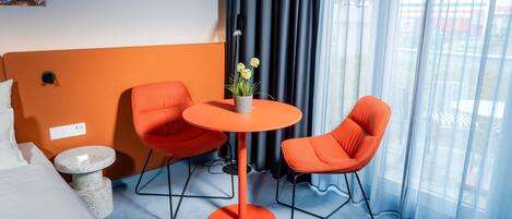 A hotel room with a bed, two orange chairs, a small table with a plant, a painting on the wall, and a window with curtains.