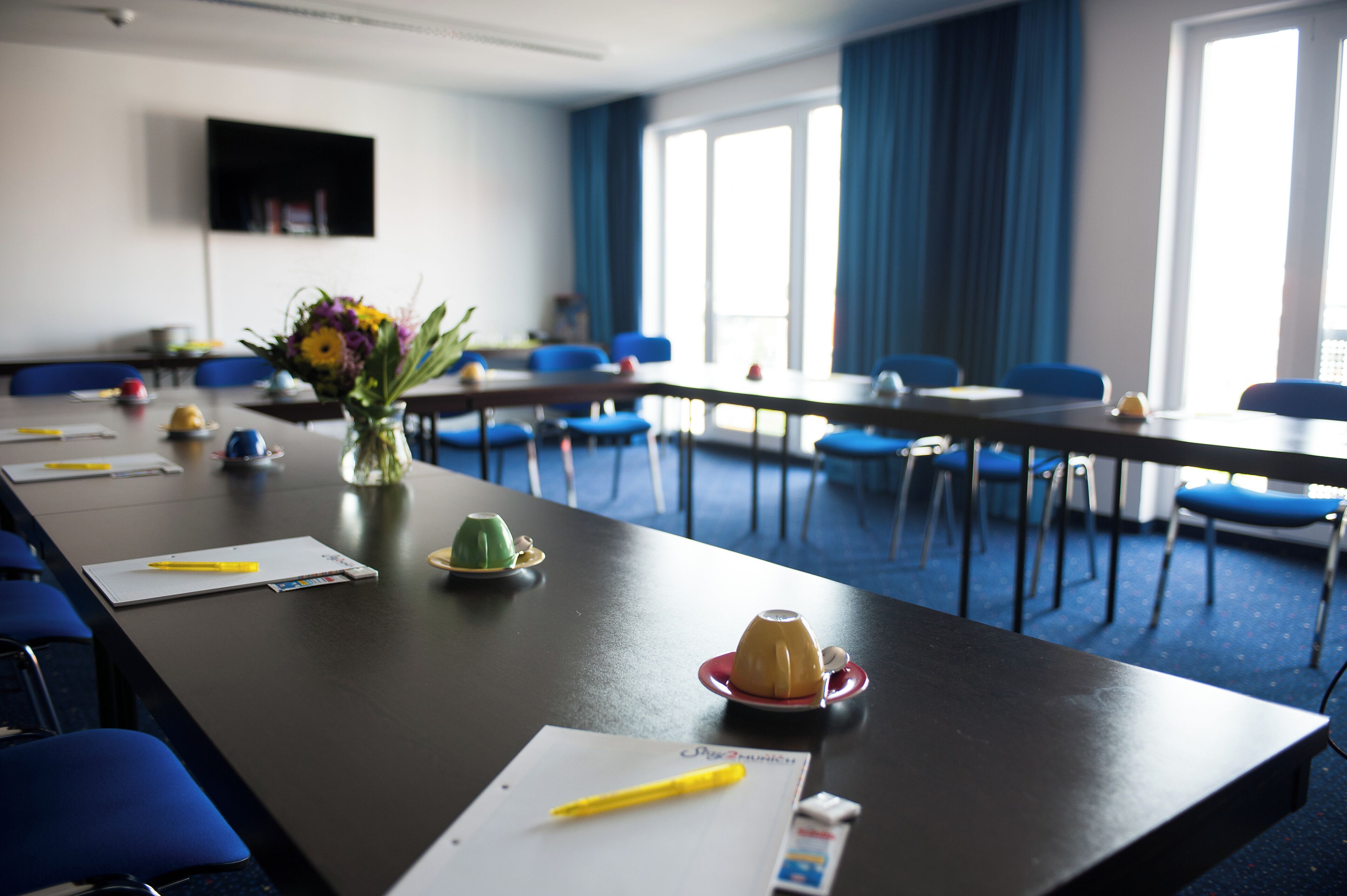 A conference room with a long table, blue chairs, a vase with flowers, and a television on the wall.