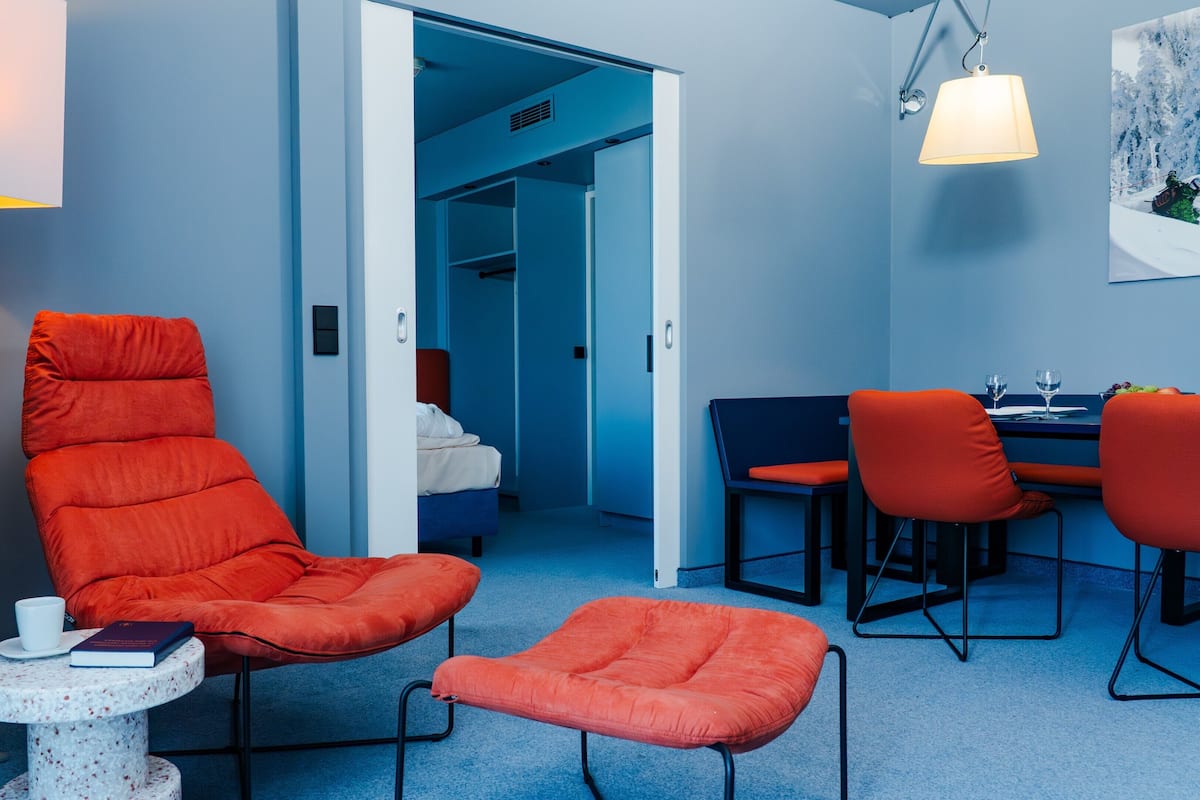 A hotel room with a red chair, a small round table with a book, a dining area with a table and chairs, and a painting on the wall.