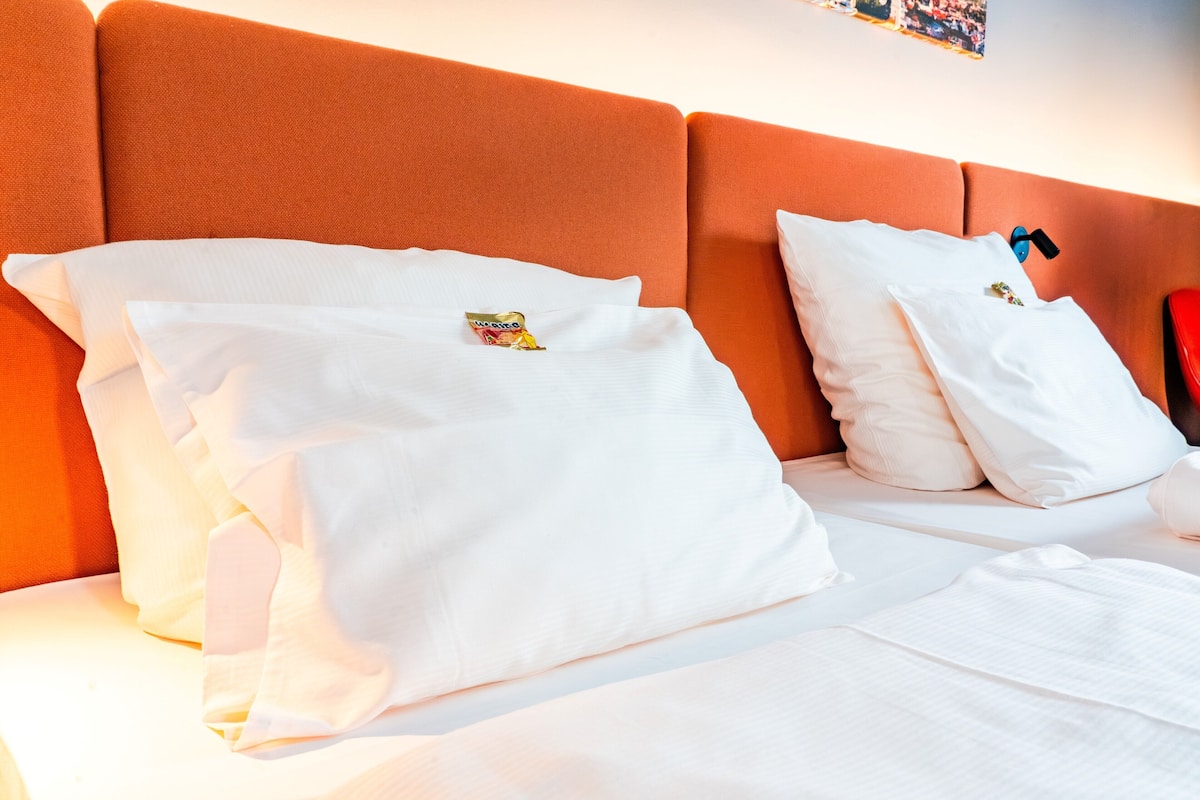 A hotel room with a large orange headboard, white bedding, and a small decorative item on the pillow.