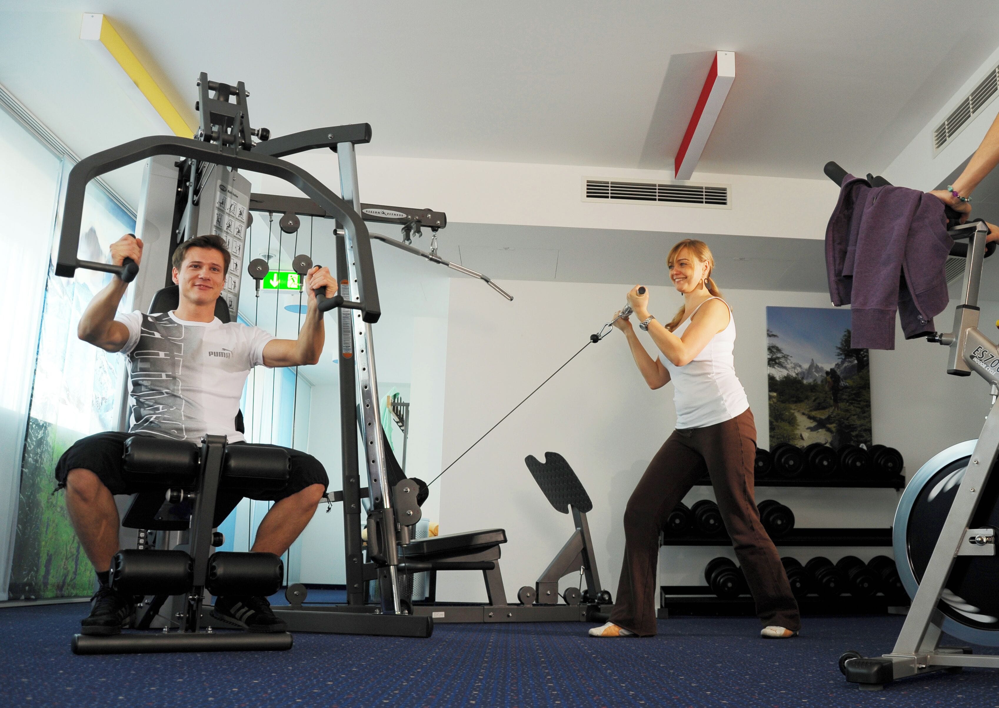A gym with exercise equipment, a wall-mounted rack of dumbbells, and a window with a view of trees.