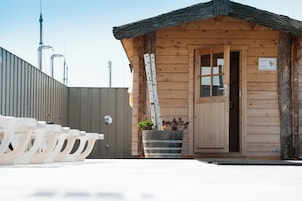 Una cabaña de madera con una puerta, una ventana y un letrero. Se ven tuberías y un árbol frente a la cabaña.