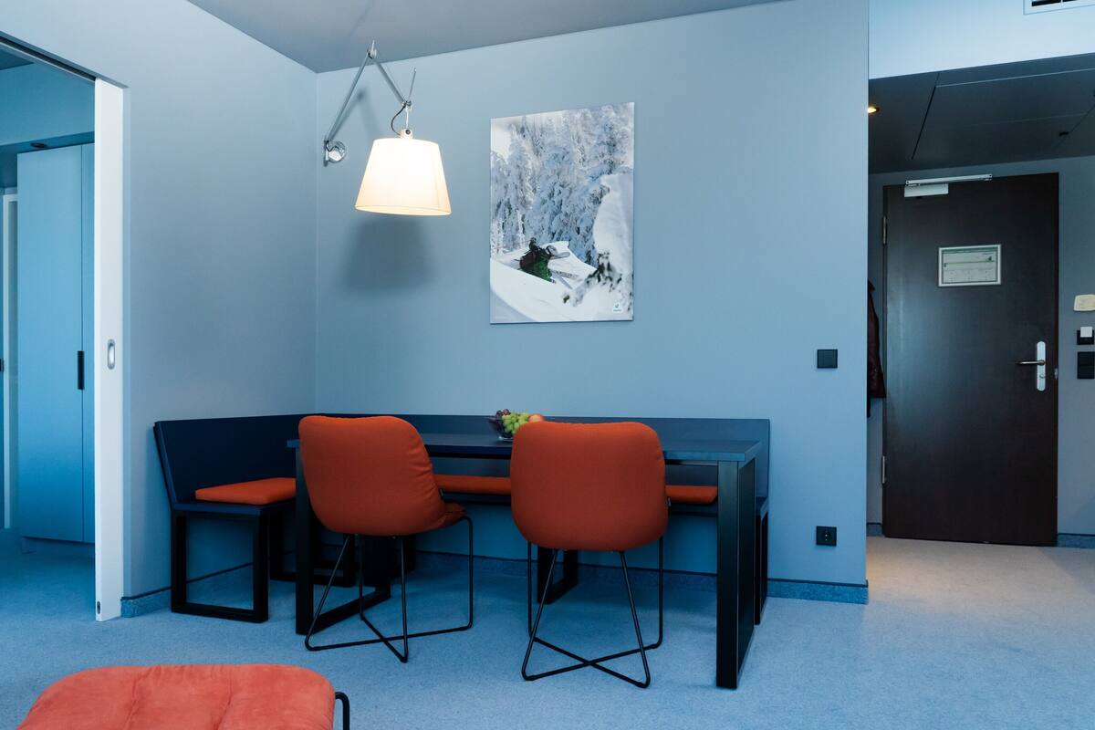 A modern hotel room with a blue wall, a dining area with a table and chairs, a bed, and a door.