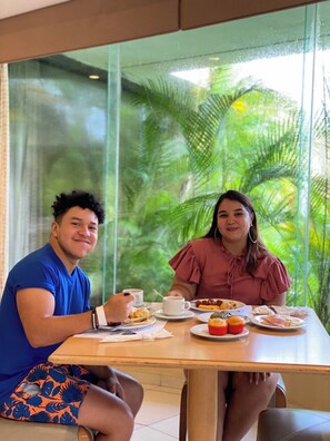 Two people seated at a table with a meal, a window with a view of greenery, and a glass partition. - Summit Rainforest Golf Resort & All Inclusive (Panama City)