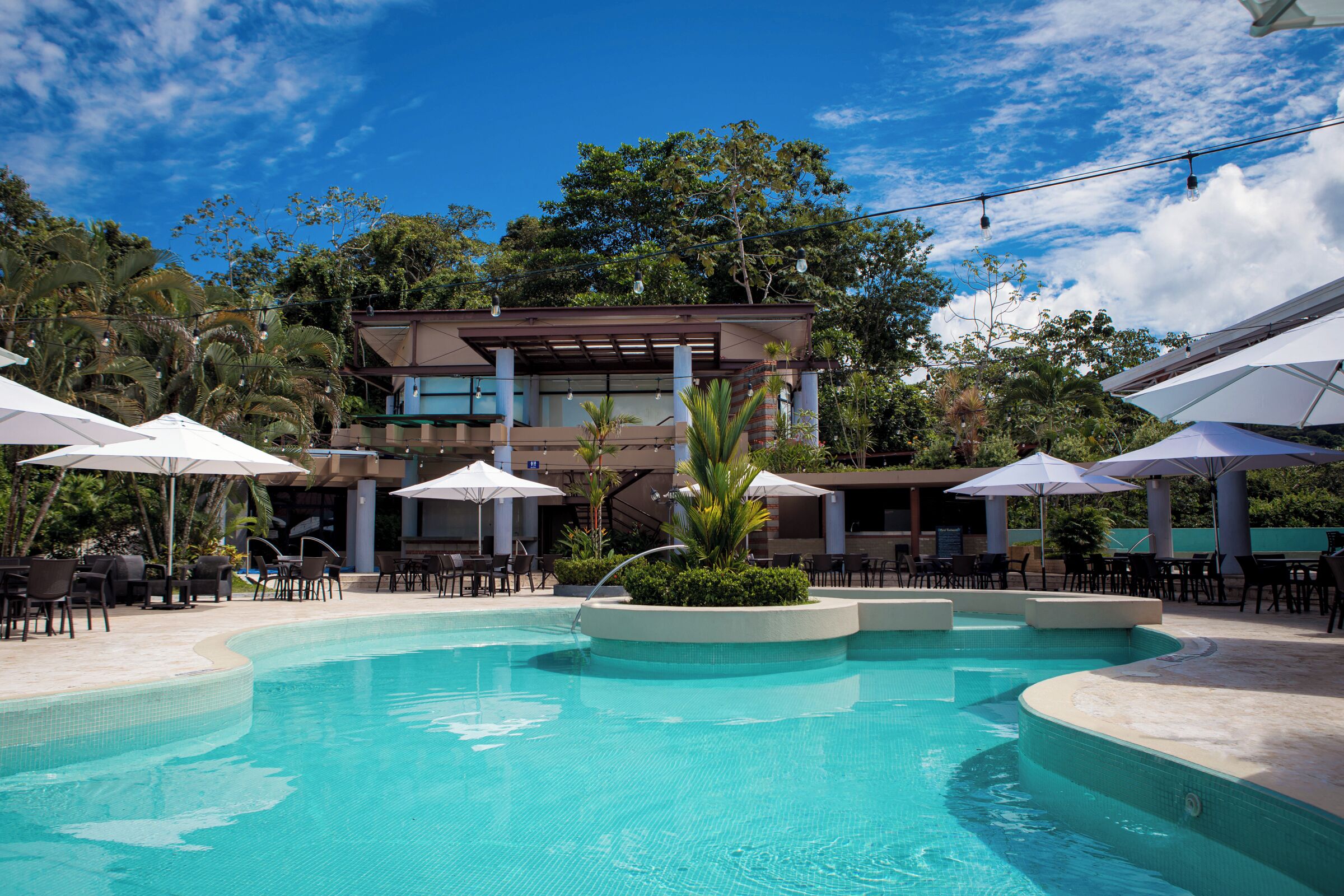 Un espace piscine doté de chaises longues et de parasols, entouré d’un bâtiment au design moderne offrant une vue sur les arbres.