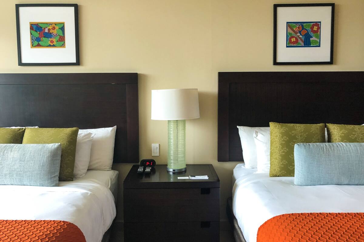 A hotel room with two beds, each with white bedding and green and blue pillows. There are framed artworks on the walls, a bedside table with a lamp, and a clock.
