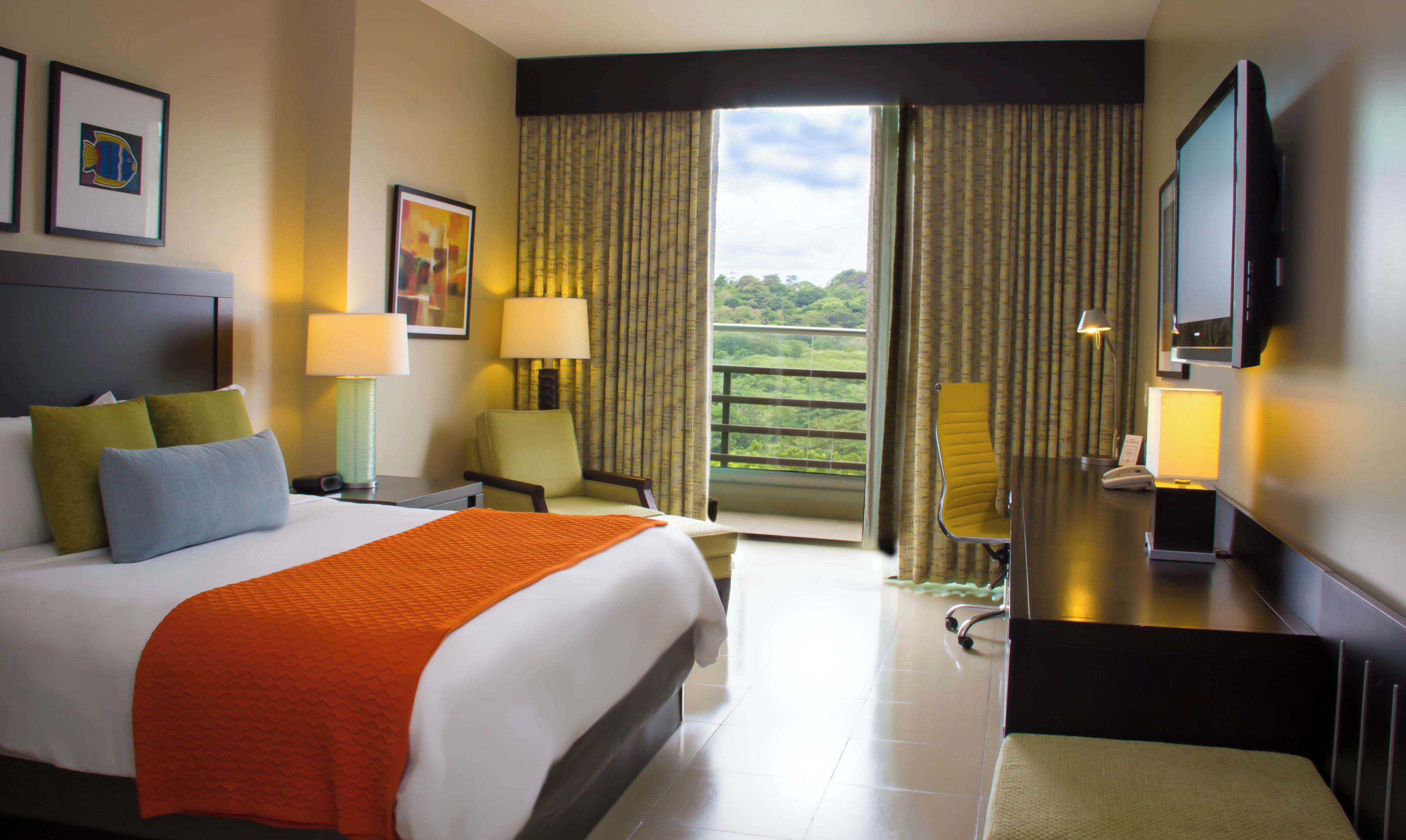 Habitación de hotel con cama, dos sillones, escritorio, televisor y balcón con cortinas.