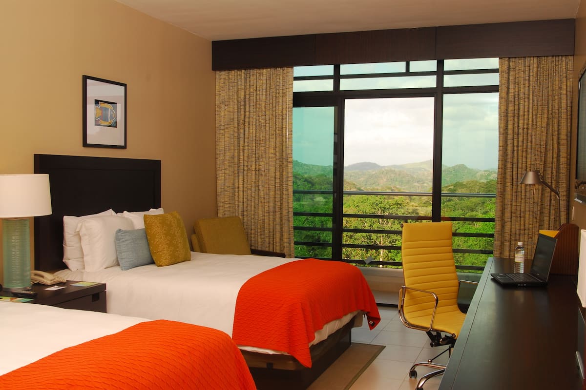 A hotel room with two beds, a desk with a laptop, a phone, and a view of greenery.
