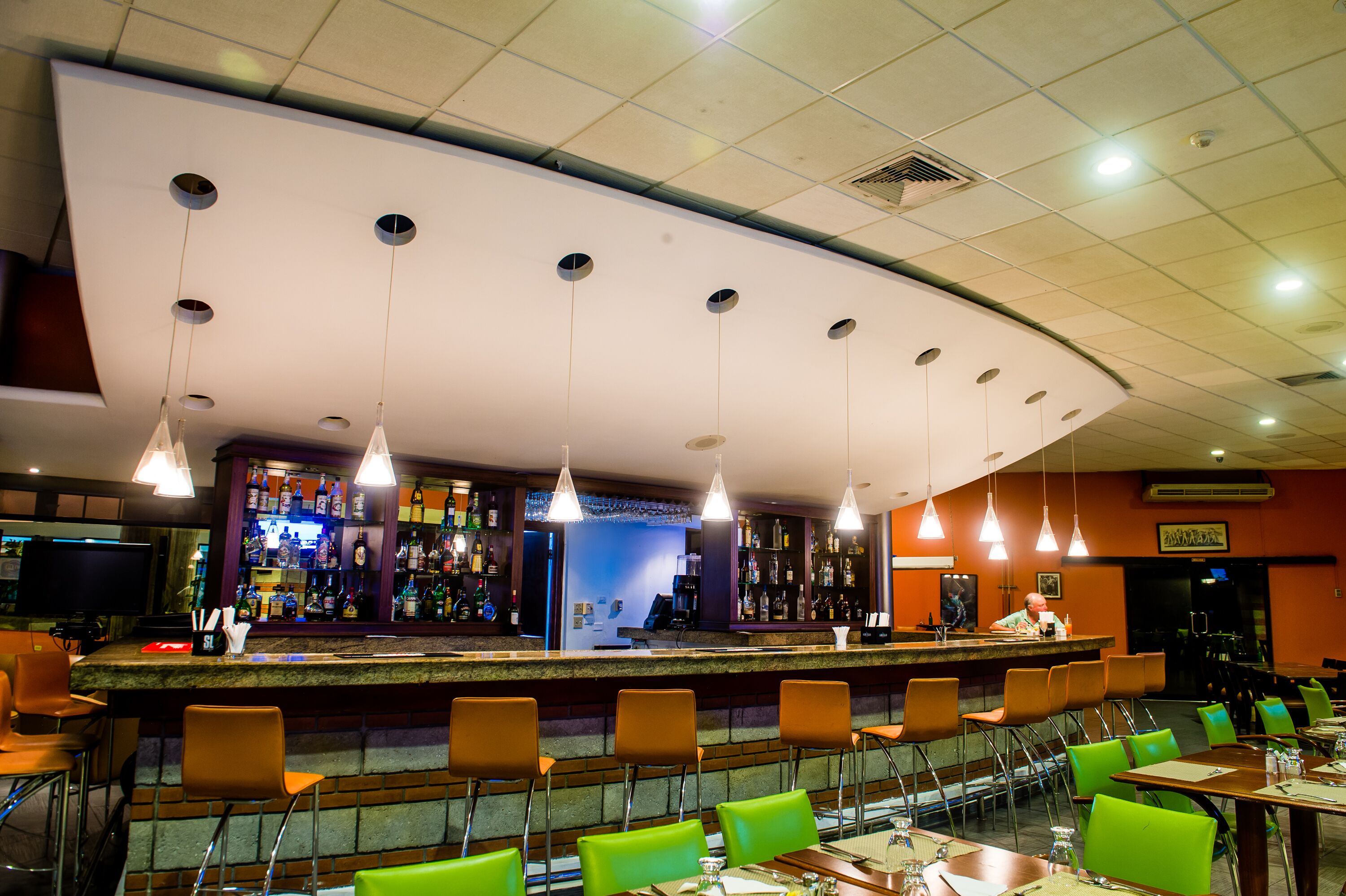Zona bar con soffitto curvo, diverse luci a sospensione e un barista. In primo piano, sgabelli da bar e tavoli con sedie.