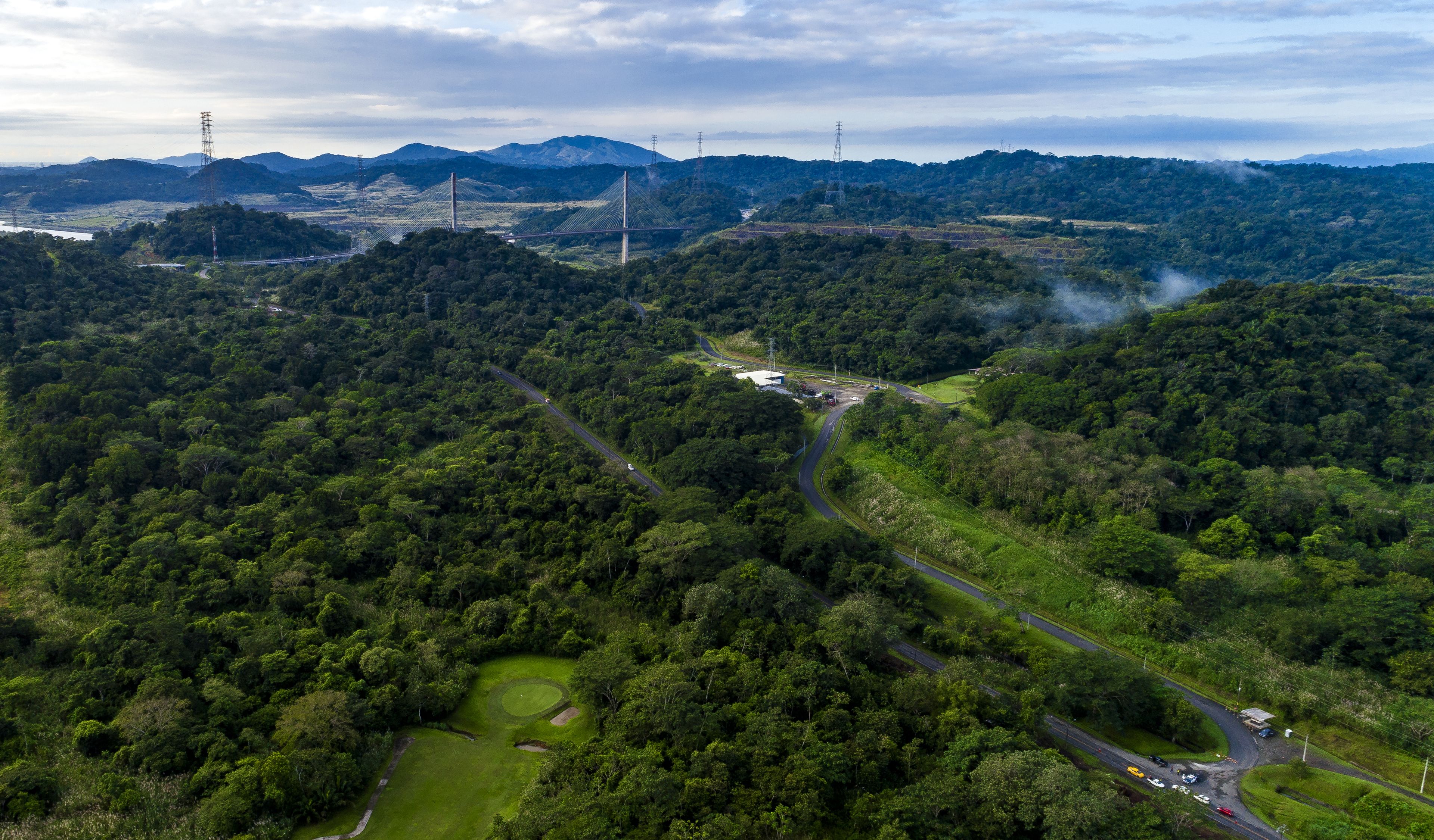 Vista aérea de un paisaje exuberante y verde con un camino sinuoso, un puente y líneas eléctricas a lo lejos.