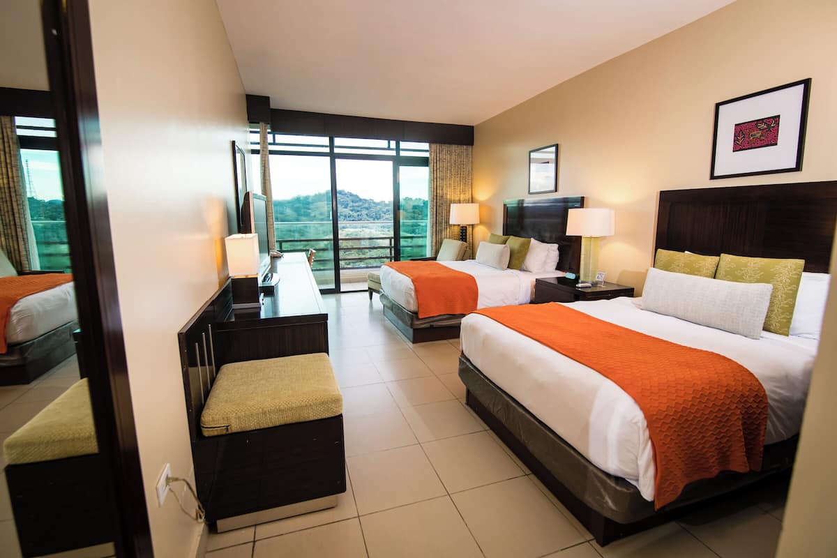 A hotel room with two beds, a desk, and a large window offering a scenic view.