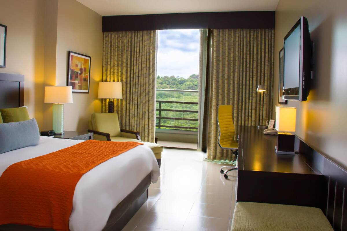 A hotel room with a bed, two chairs, a desk, a TV, and a balcony with curtains.
