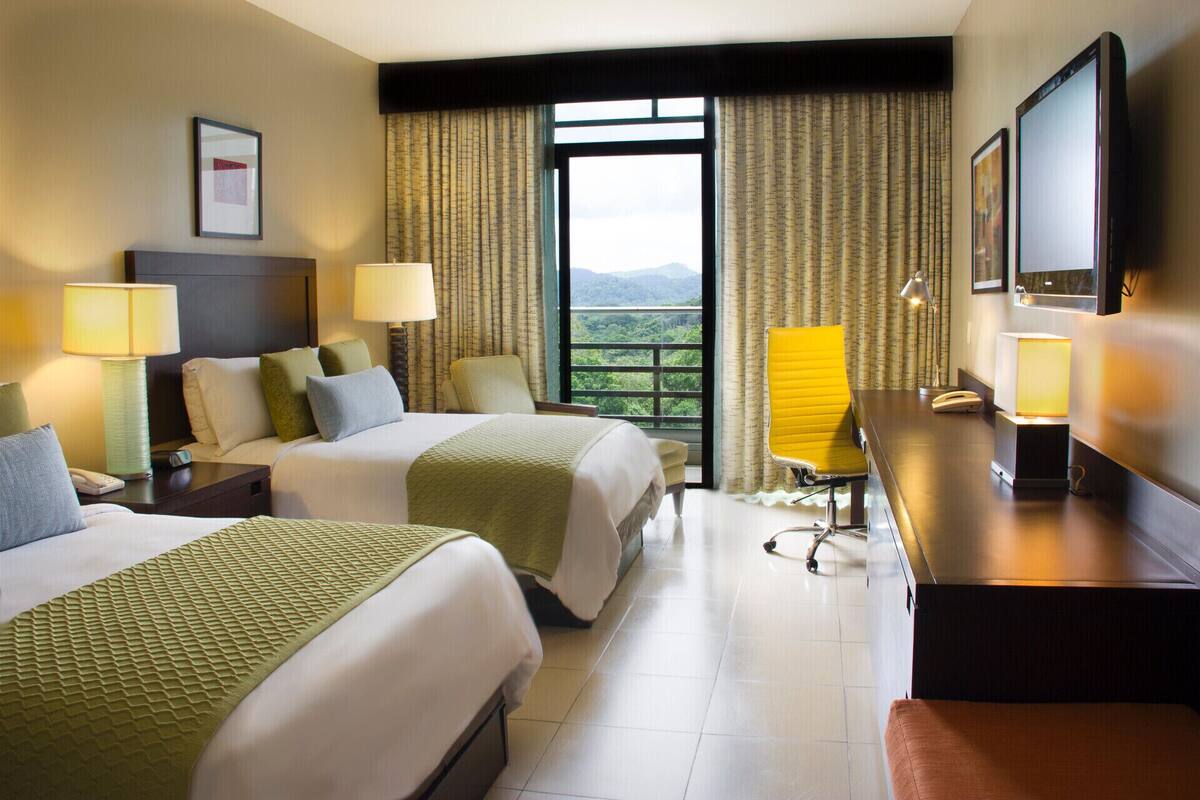 A hotel room with two beds, a desk, a chair, a TV, and a window with a view.