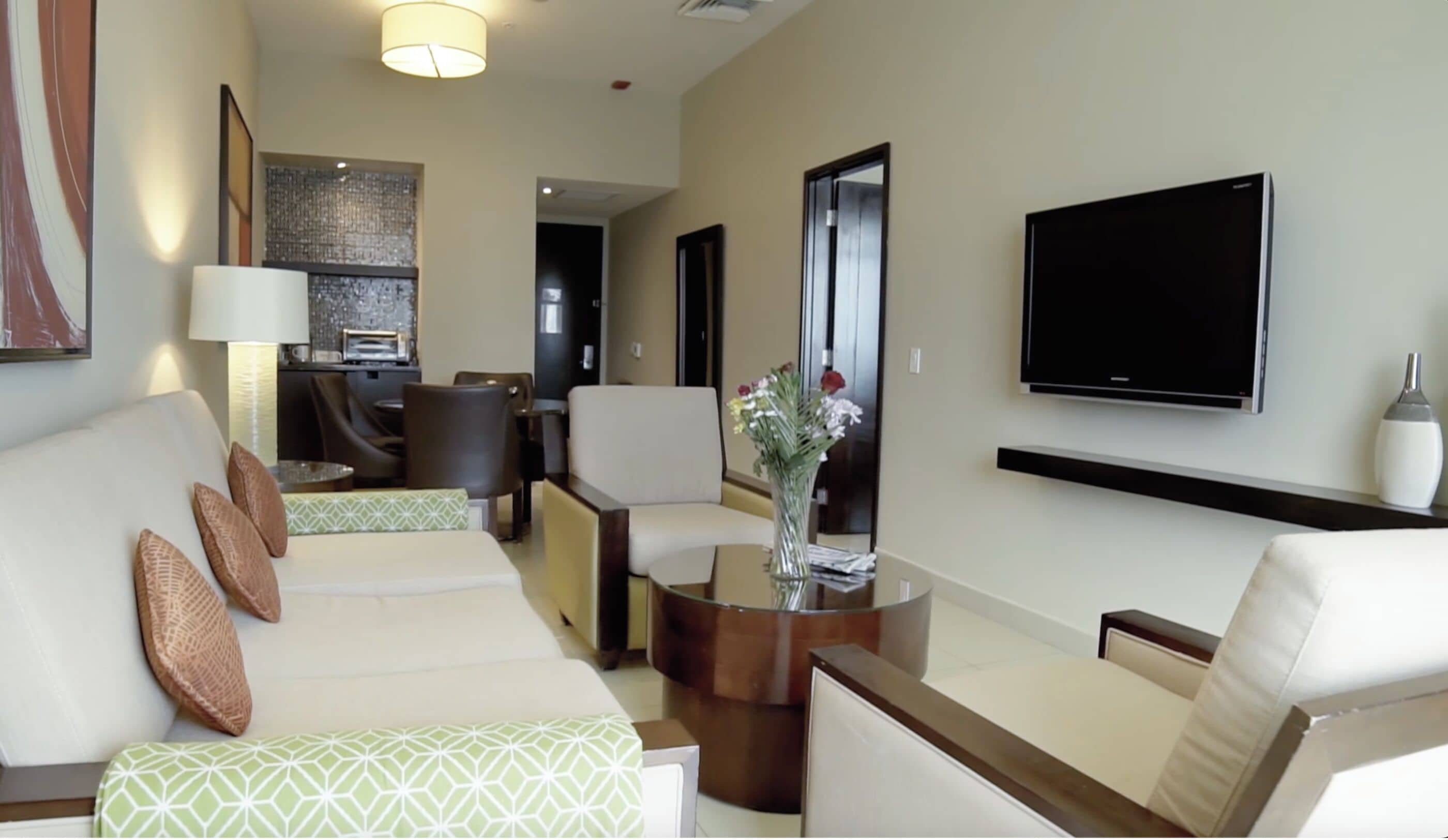 Una habitación de hotel moderna con sofá, sillones, una mesita de centro, televisor de pantalla plana y un pequeño rincón de cocina.