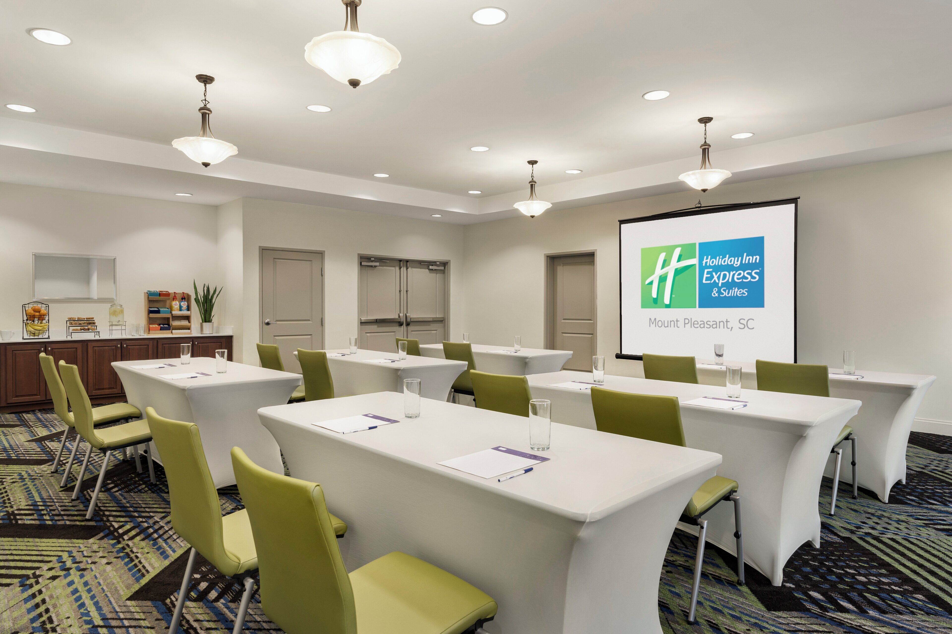 Un salón de conferencias con mesas blancas, sillas verdes y una pantalla de proyección que muestra el logotipo de Holiday Inn Express.