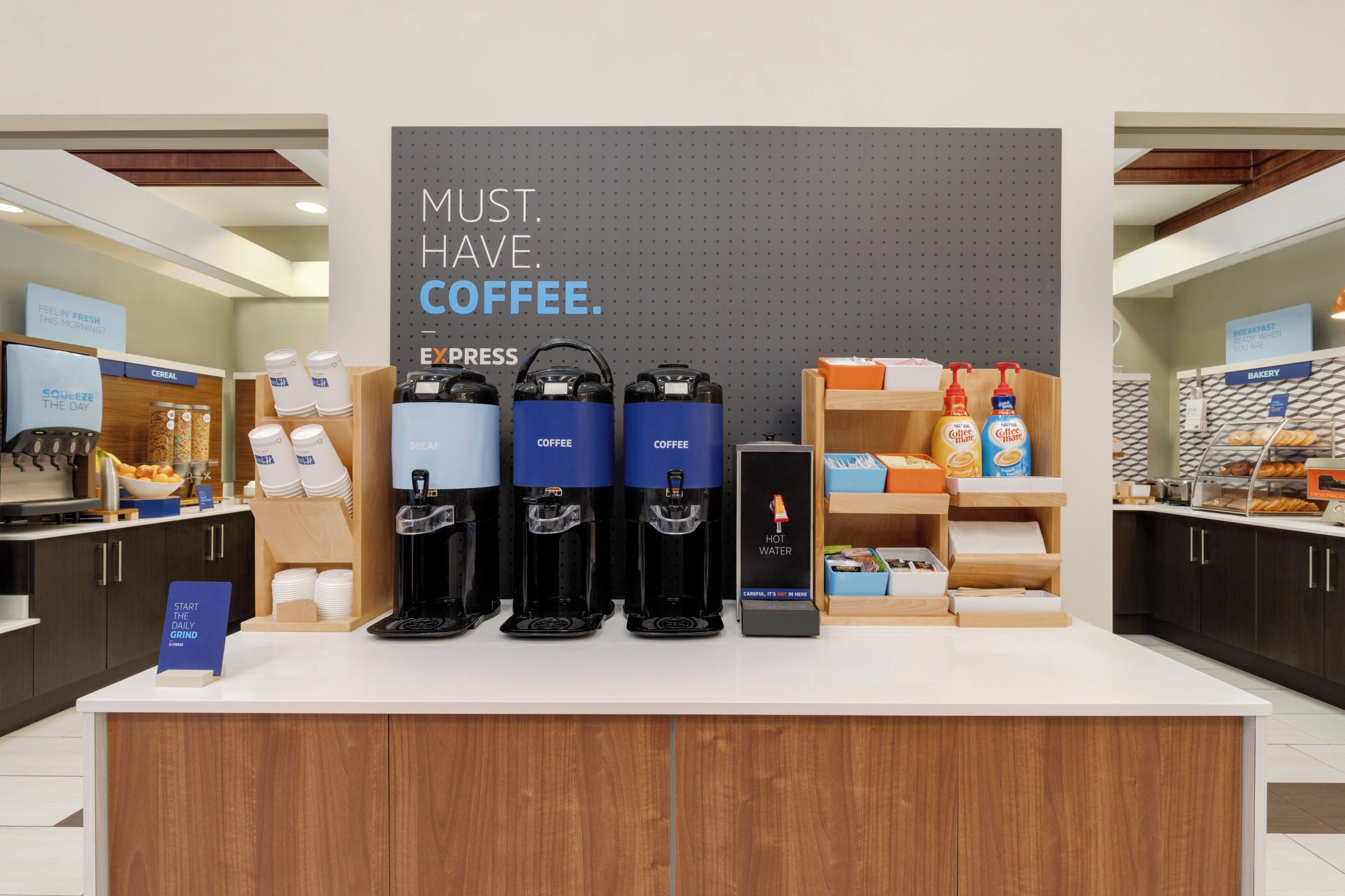 Una estación de café con varios dispensadores, un cartel que dice 'MUST. HAVE. COFFEE.' y diversas opciones para el desayuno.