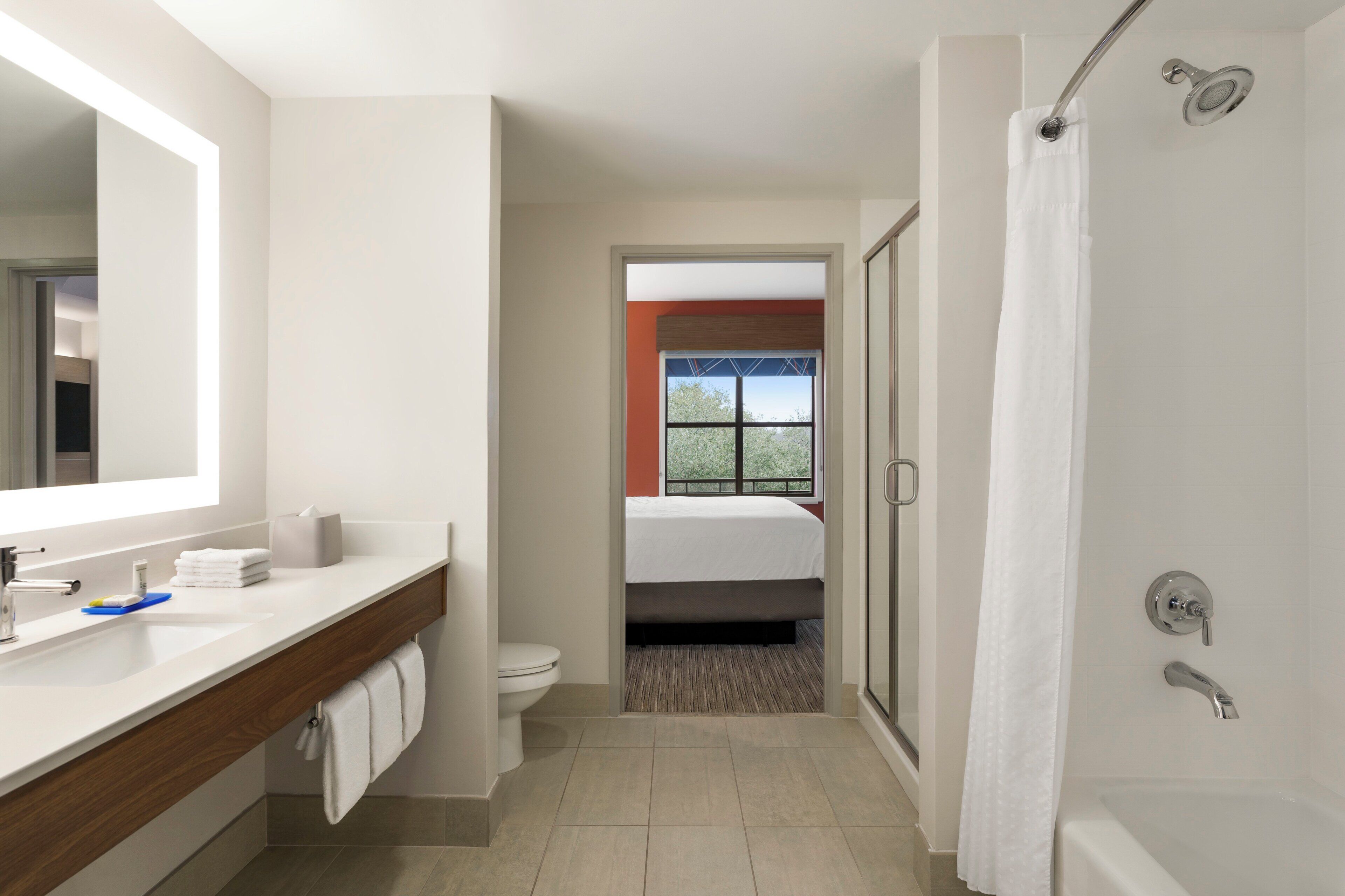 Een moderne badkamer met een douche, toilet en een bed dat door een geopende deur zichtbaar is.