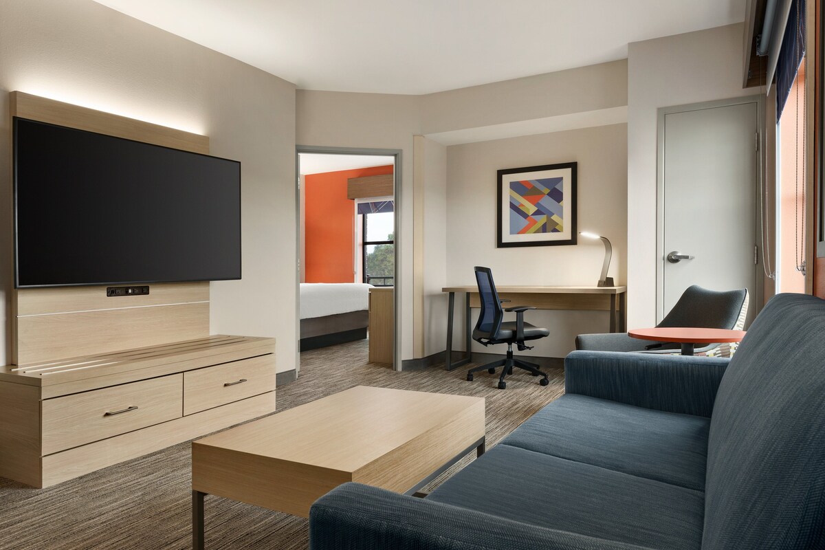 A modern hotel room with a flat-screen TV, a wooden dresser, a blue sofa, a desk with a chair, and a painting on the wall.