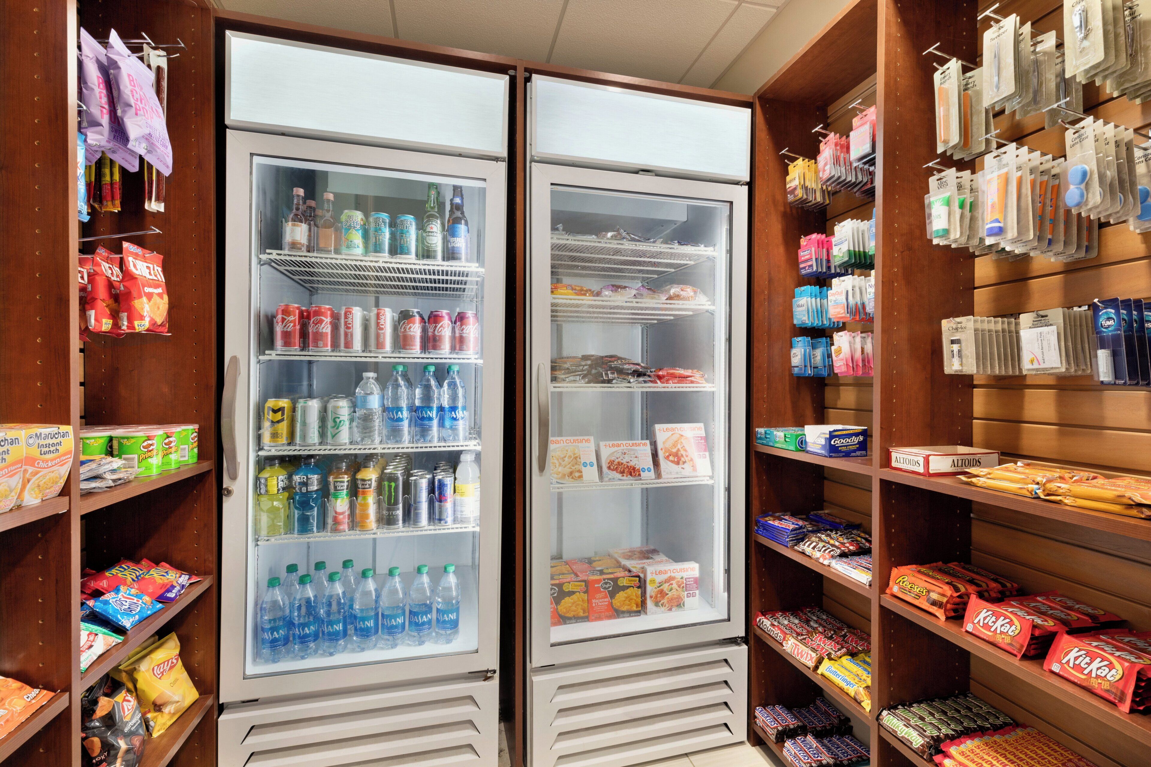 A hotel mini-fridge stocked with beverages and snacks, adjacent to shelves with various packaged goods.