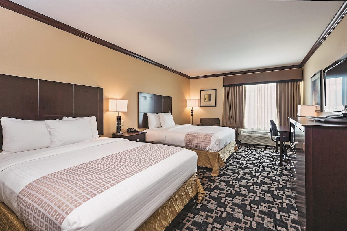 A hotel room with two beds, a desk, a chair, and a large window with curtains.