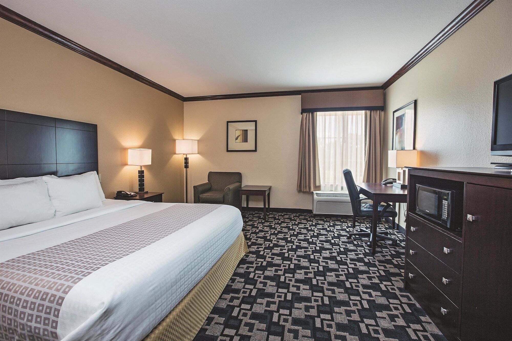 A hotel room with a large bed, two bedside lamps, a desk with a chair, a television, and a patterned carpet.