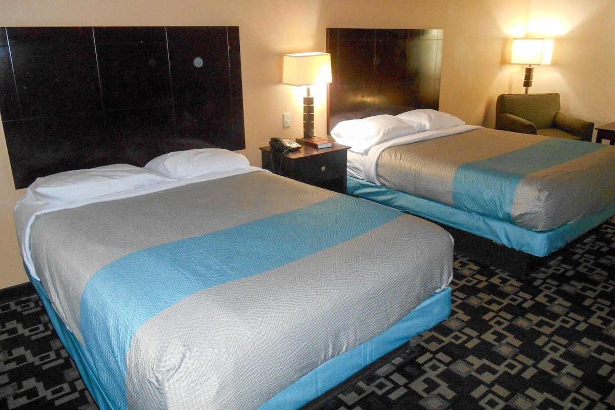 A hotel room with two beds, a nightstand with a lamp, and a chair.