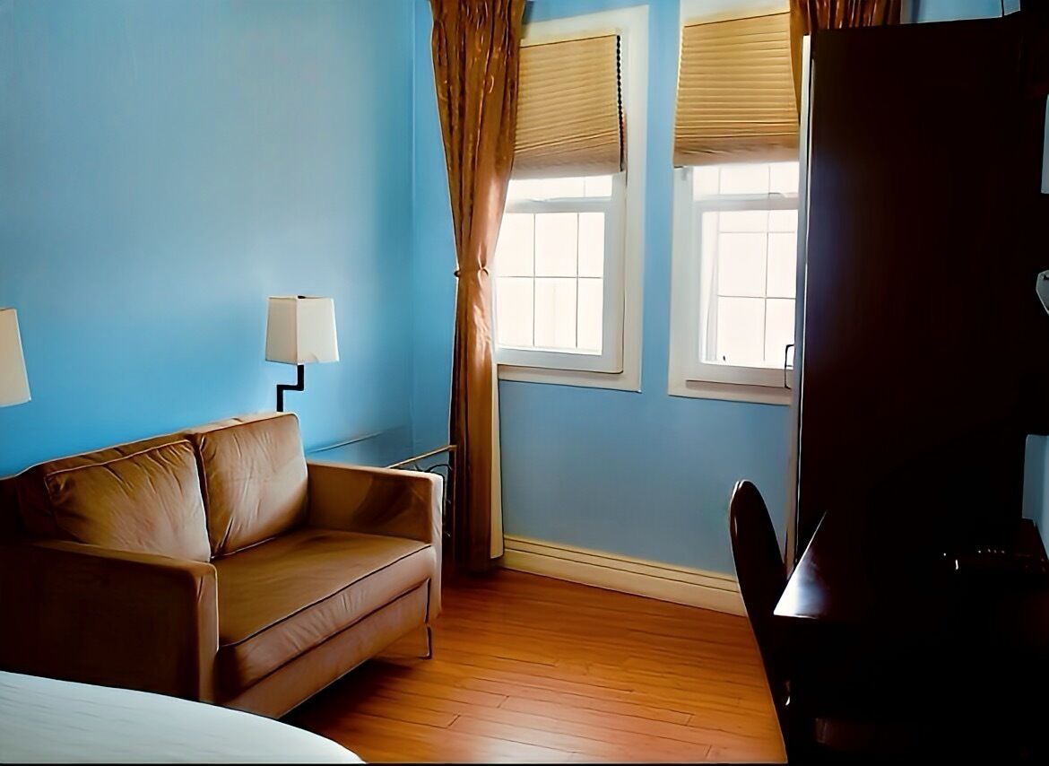 A room with a brown leather sofa, a desk with a lamp, two windows with blinds, and a dark-colored wardrobe.