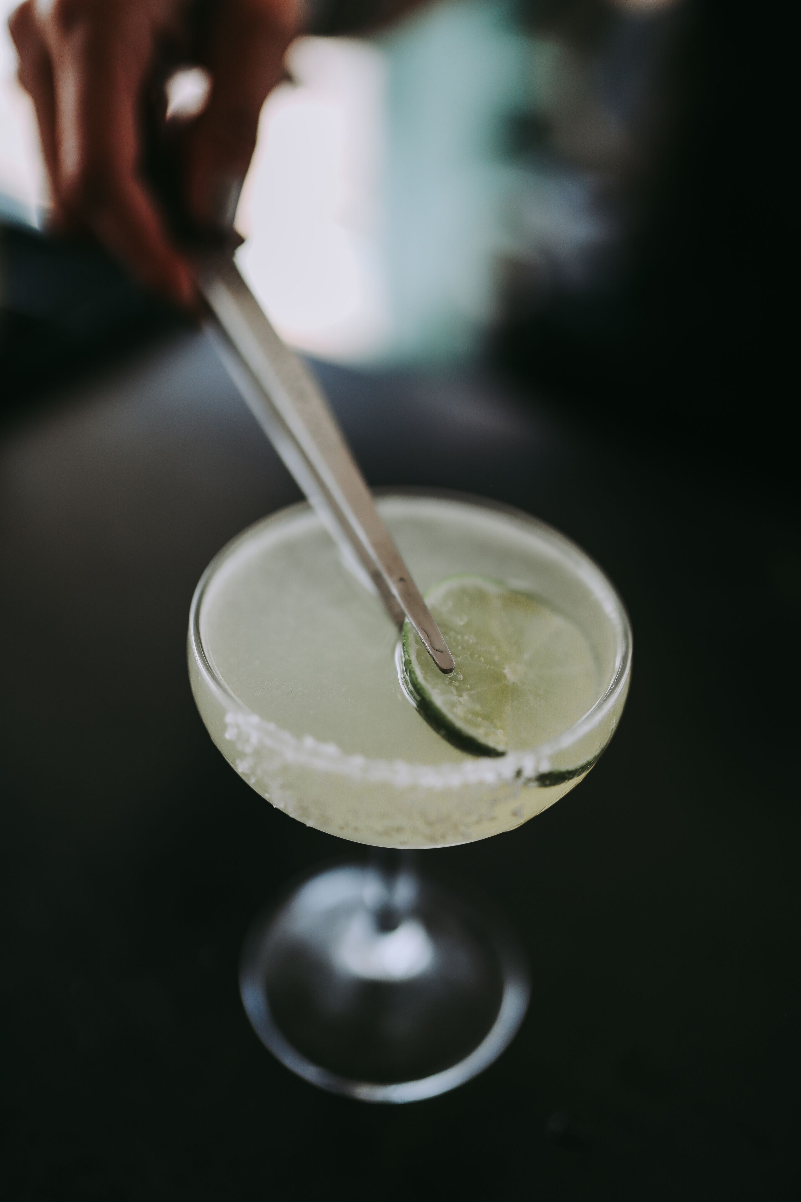 A cocktail glass with a lime wedge on the rim, a spoon inside, and a blurred background.