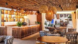 A spacious lounge area with a high ceiling made of natural materials, a central wooden table surrounded by wicker chairs, and a bar area with shelves of bottles and a plant. - Sauipe Brisa Grand Premium – All Inclusive (Mata de São João)