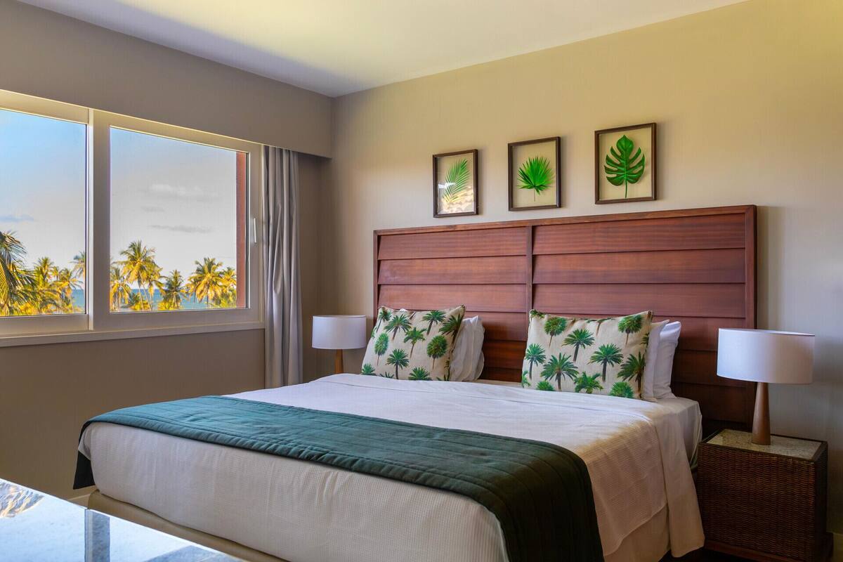 A hotel room with a bed, a window with a view of palm trees, and three framed pictures on the wall.