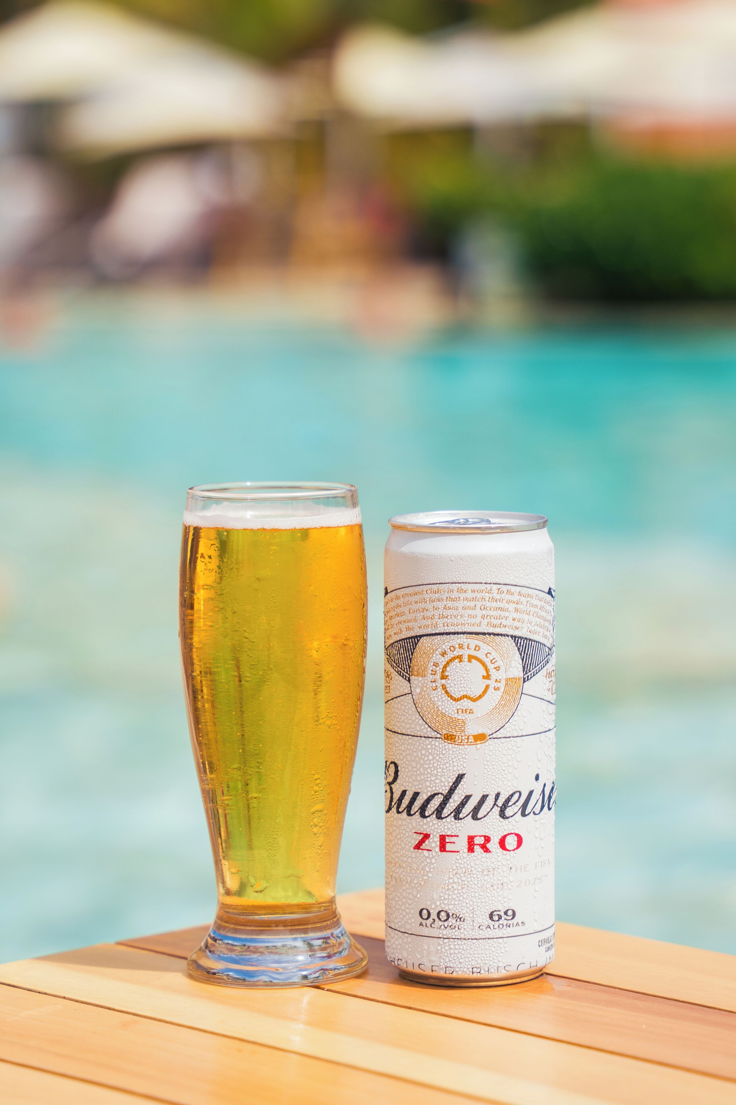A glass of beer next to a Budweiser Zero can on a wooden table by a pool.