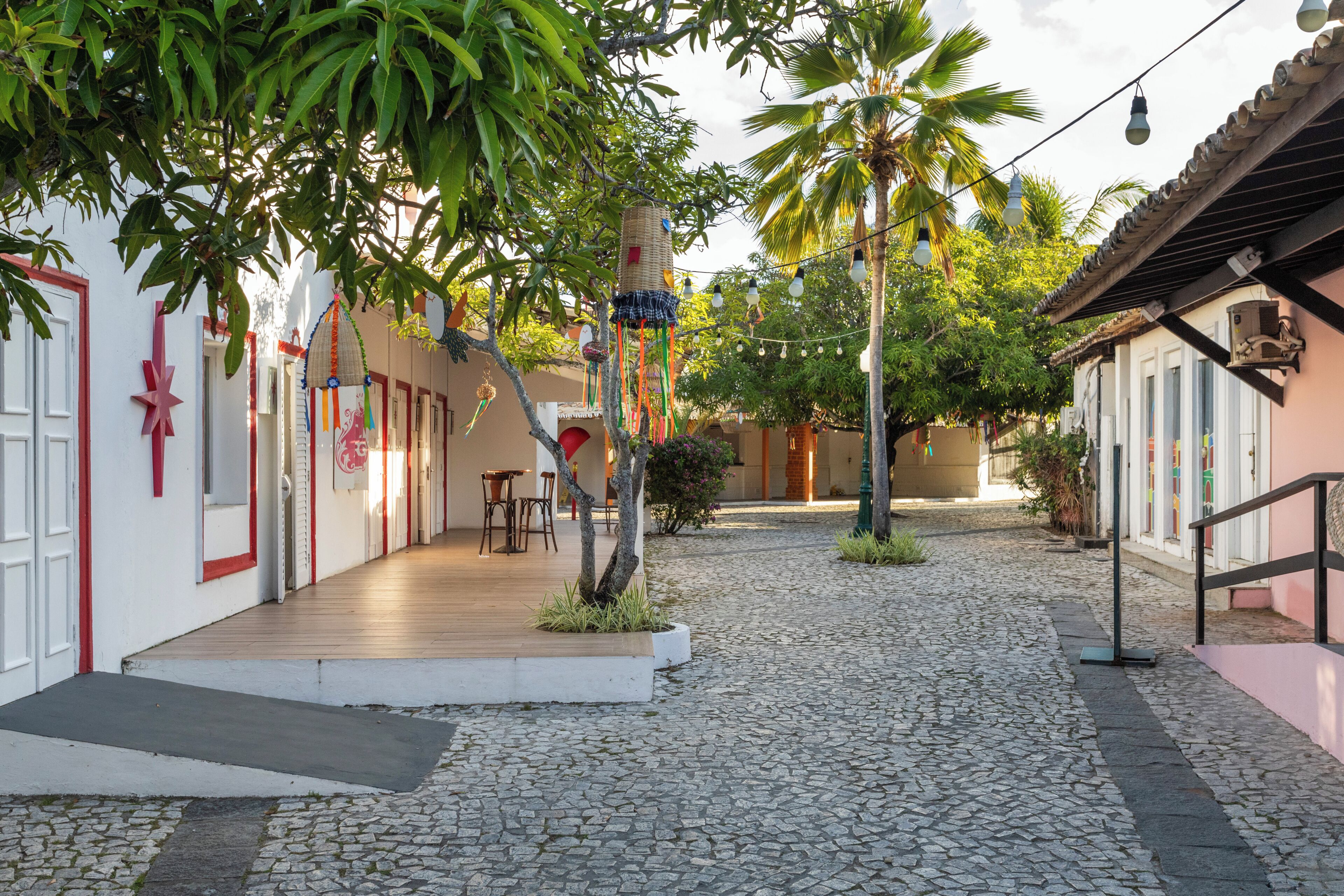 Un patio empedrado con una terraza de madera, palmeras y edificios con detalles blancos y rojos.