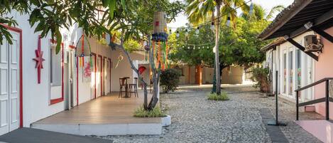 Un patio empedrado con una terraza de madera, palmeras y edificios con detalles blancos y rojos.
