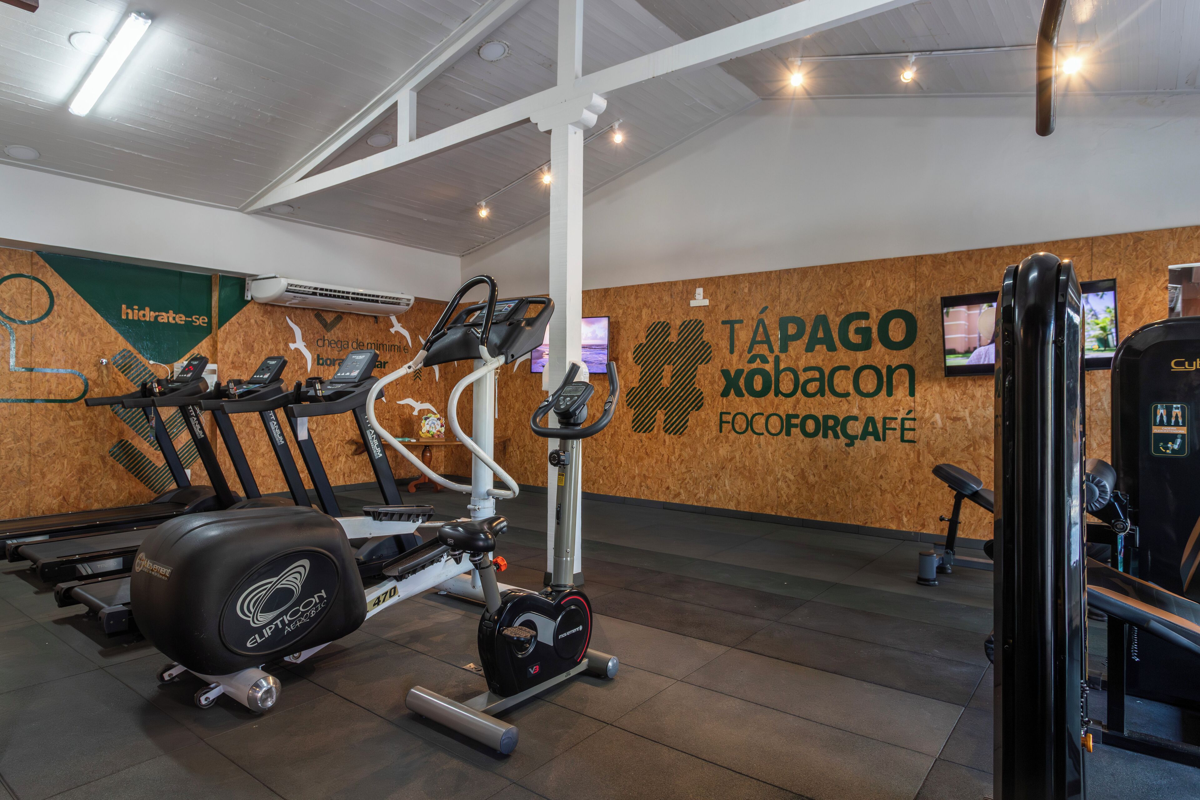 A gym with exercise equipment, a wooden wall with a logo, and a ceiling with lights.