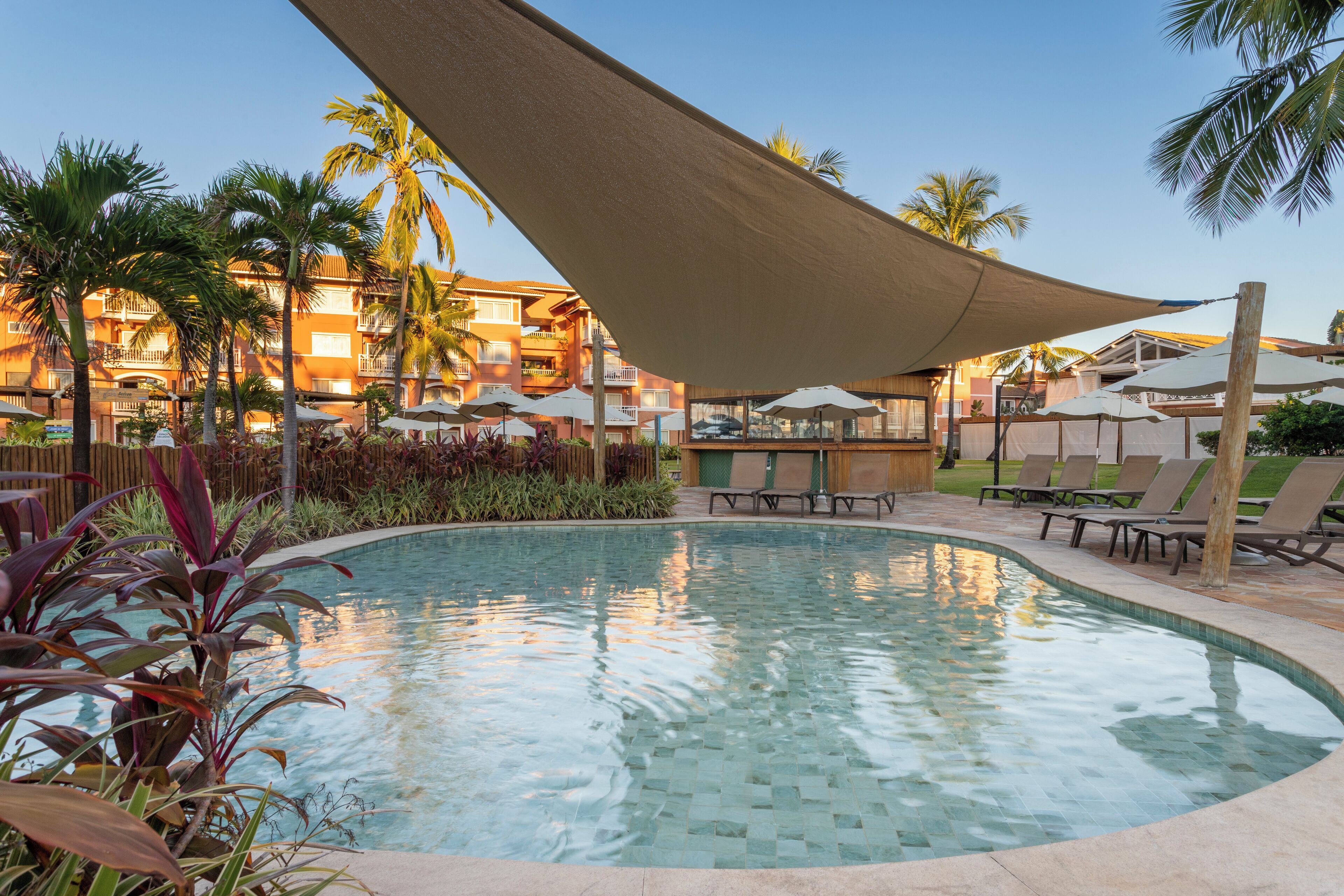 A swimming pool with lounge chairs and umbrellas, surrounded by palm trees and a building with balconies.