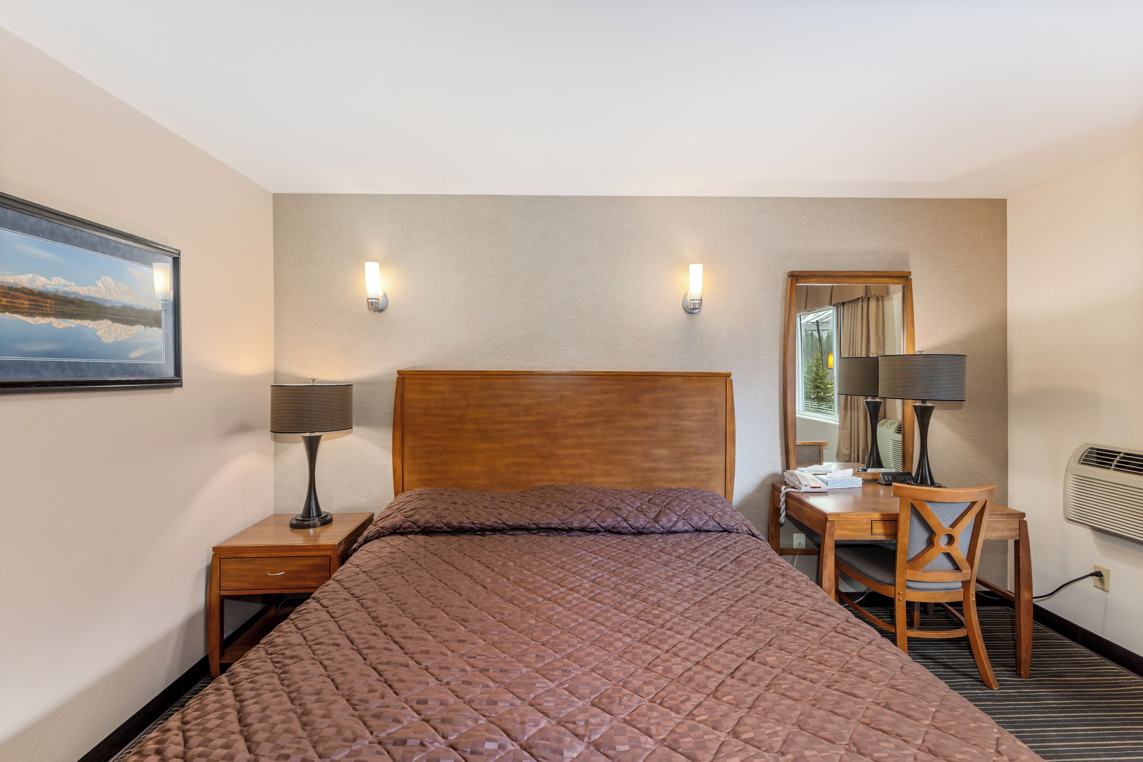 A hotel room with a large bed, a wooden nightstand, a desk with a chair, a lamp, and a mirror.